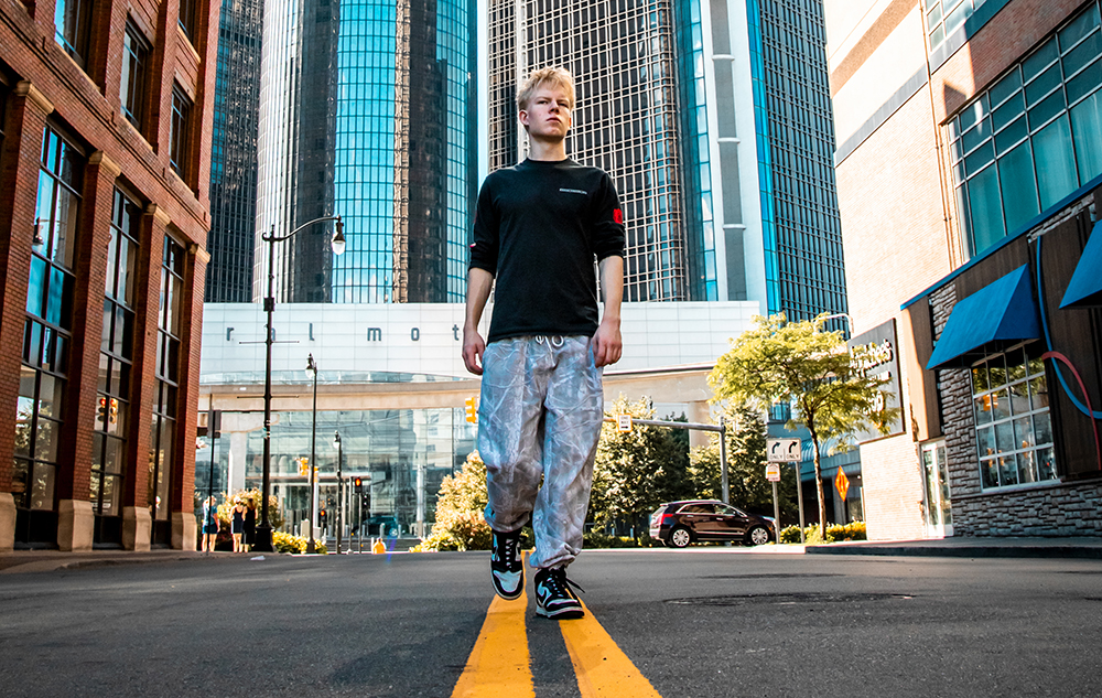 A high school senior guy walks down the center of a city street between tall buildings, with the Renaissance Center skyscrapers and a bright blue sky in the background, capturing a memorable moment from his Novi senior pictures session.