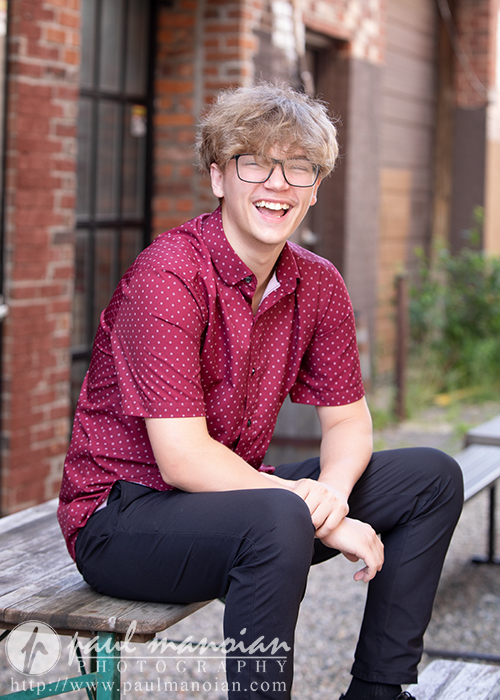 A smiling high school senior with glasses and tousled hair sits on a bench outside, wearing a red patterned shirt and black pants. Brick buildings and greenery are visible in the background.