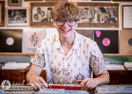 A high school senior browses through vinyl records for their senior pictures session in a cozy, vintage record store filled with framed photos.