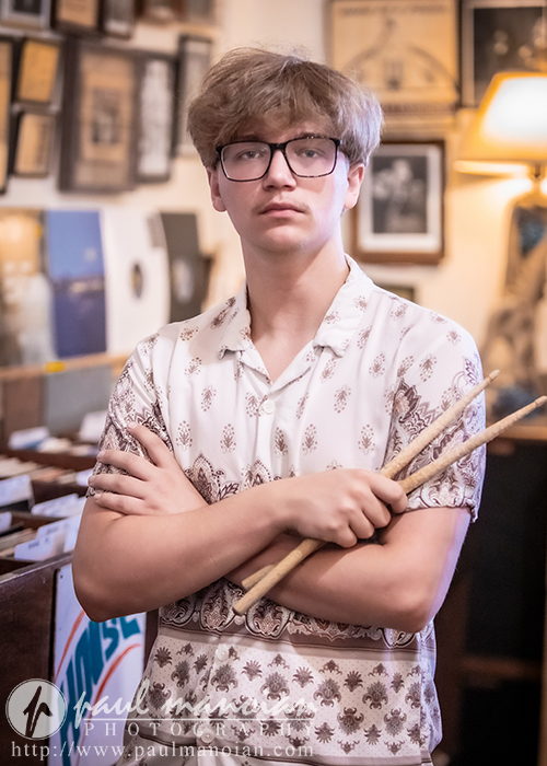 A young person with glasses and a patterned shirt stands indoors, holding drumsticks with arms crossed. The background includes framed pictures, a lamp, and shelves, creating a cozy, artistic atmosphere.