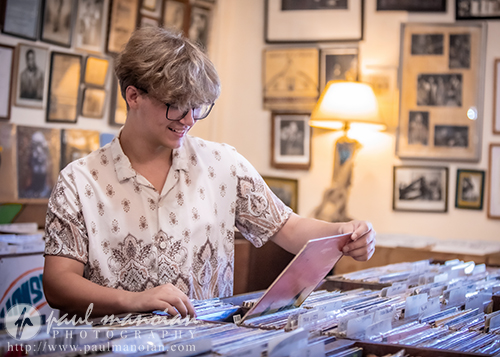 A high school senior browses through vinyl records for their senior pictures session in a cozy, vintage record store filled with framed photos.
