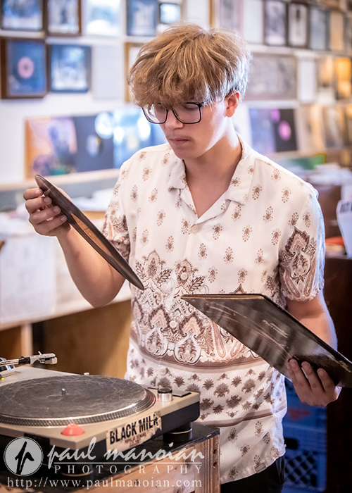 A young person with glasses and messy hair examines a vinyl record near a turntable inside a record store. Framed albums decorate the wall in the background.