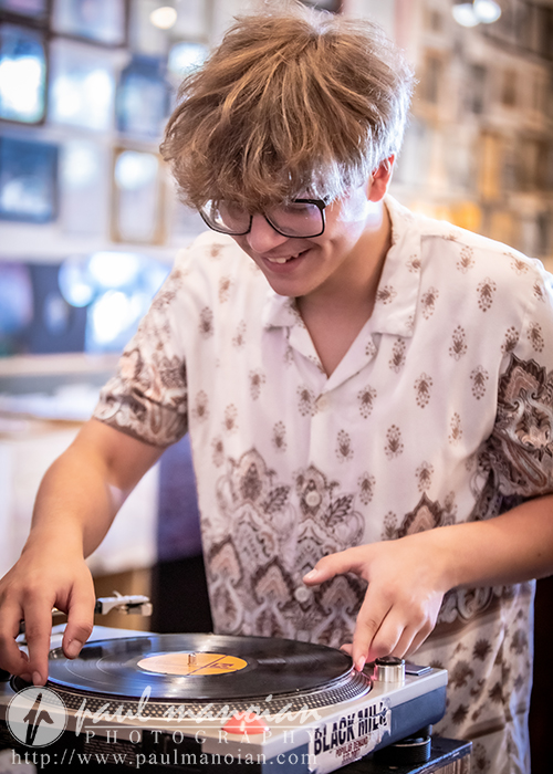 A young person with glasses and tousled hair smiles while operating a vinyl record player, wearing a patterned button-up shirt in a cozy indoor setting with blurred photos on the wall behind them.