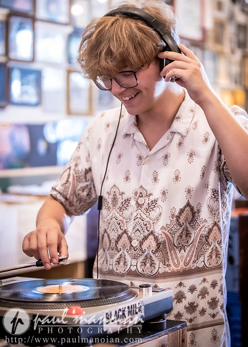 A young person with shaggy hair and glasses smiles while using headphones and adjusting a vinyl record on a turntable in a cozy, art-filled room.