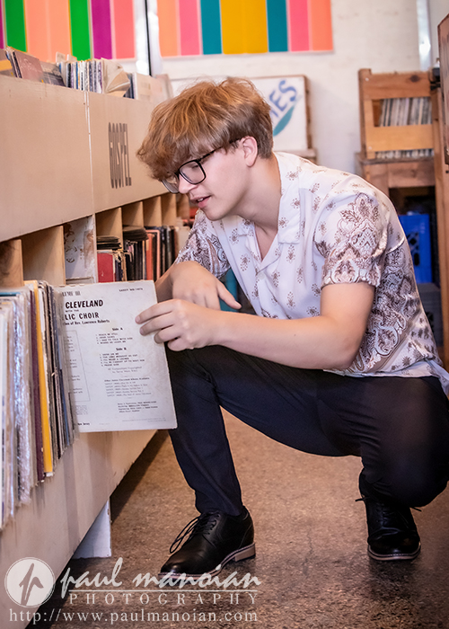 A young man with glasses and a patterned shirt kneels in a record store, browsing vinyl records. He holds a Cleveland Public Choir album, surrounded by shelves of records. The store has a colorful, artistic atmosphere.