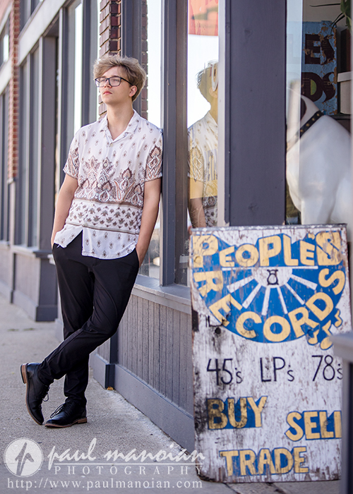 A high school senior in glasses and a patterned shirt leans against a storefront window next to a weathered sign that reads "People's Records: 45s LP's 78's BUY SELL TRADE" on a city sidewalk.