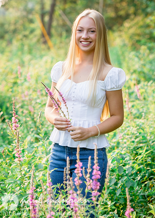 A young woman with long blond hair, wearing a white blouse and blue jeans, stands in a sunlit field of wildflowers during her Ann Arbor senior pictures session, smiling and holding purple flowers amid green foliage and pink blossoms.