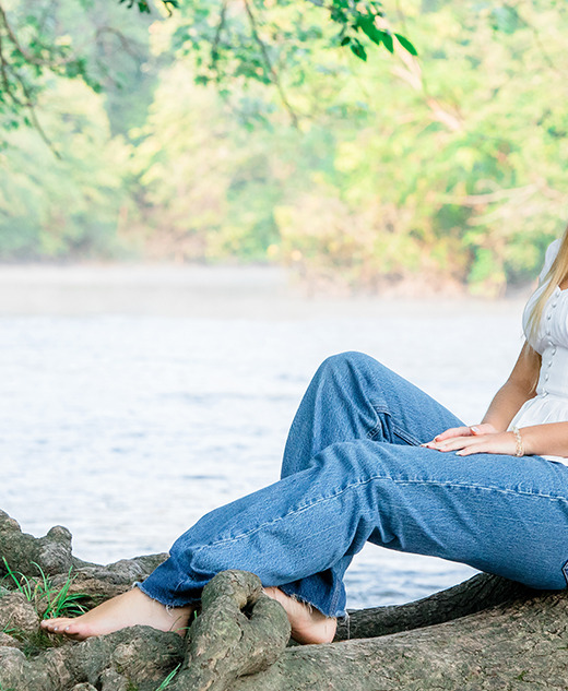 A young woman with long blonde hair, wearing a white top and blue jeans, sits barefoot against a large tree by a river during her Ann Arbor senior pictures session, smiling at the camera. Lush green foliage and water are visible in the background.
