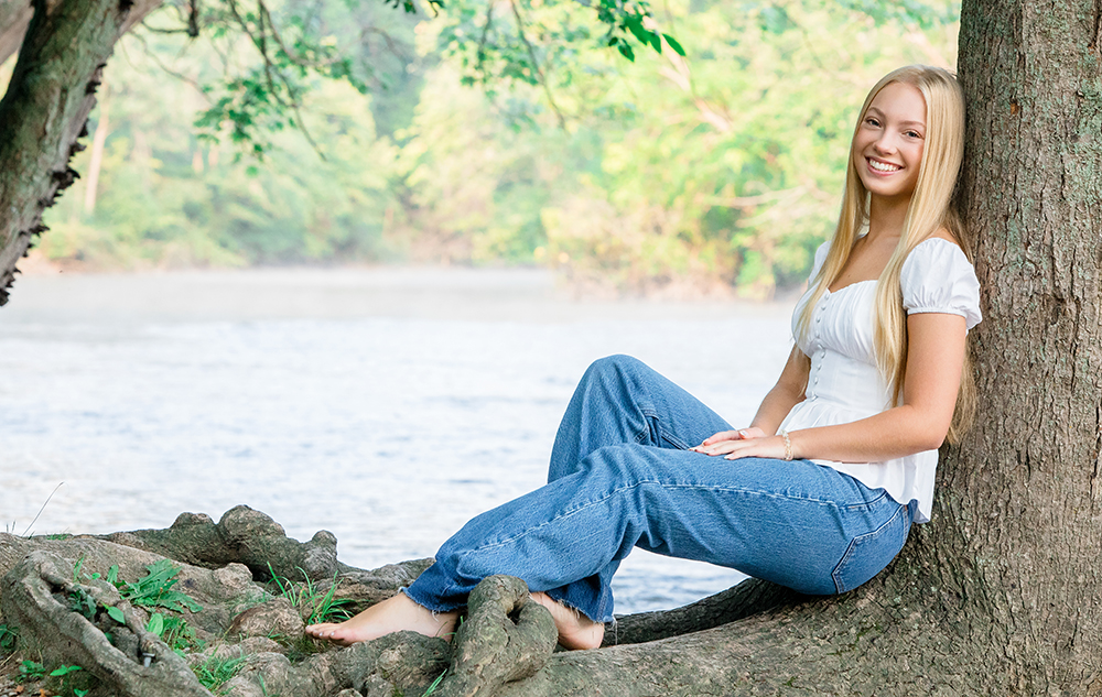 A young woman with long blonde hair, wearing a white top and blue jeans, sits barefoot against a large tree by a river during her Ann Arbor senior pictures session, smiling at the camera. Lush green foliage and water are visible in the background.