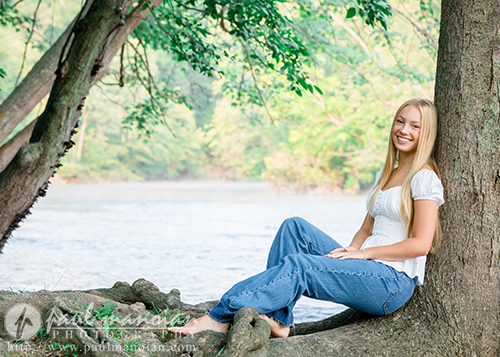 A young woman with long blonde hair, wearing a white top and blue jeans, sits barefoot against a tree by a river, smiling at the camera—lush green foliage surrounds her during this Ann Arbor senior pictures session.