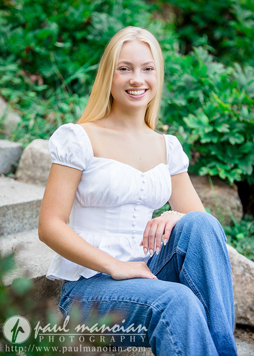 A young woman with long blonde hair, wearing a white blouse and blue jeans, sits on outdoor stone steps surrounded by greenery, smiling at the camera during her Ann Arbor senior pictures session.