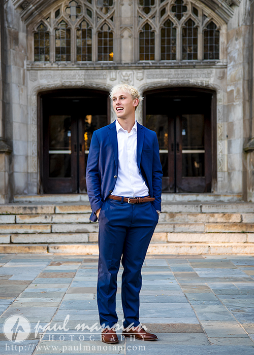 A young man with blond hair in a blue suit and white shirt stands smiling in front of a large, ornate stone building with steps and arched windows during his Ann Arbor senior portraits session.