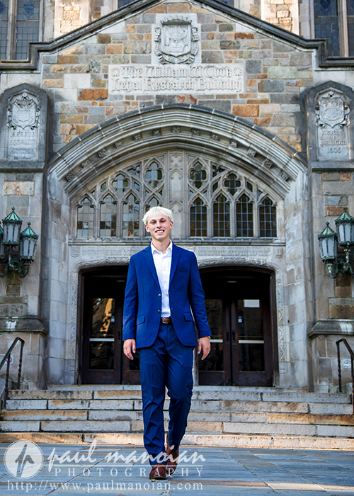 A young man in a blue suit stands smiling on the steps outside a stone university building with arched windows and ornate lamps, capturing a memorable Ann Arbor senior portraits session. The building's name is engraved above the entrance.