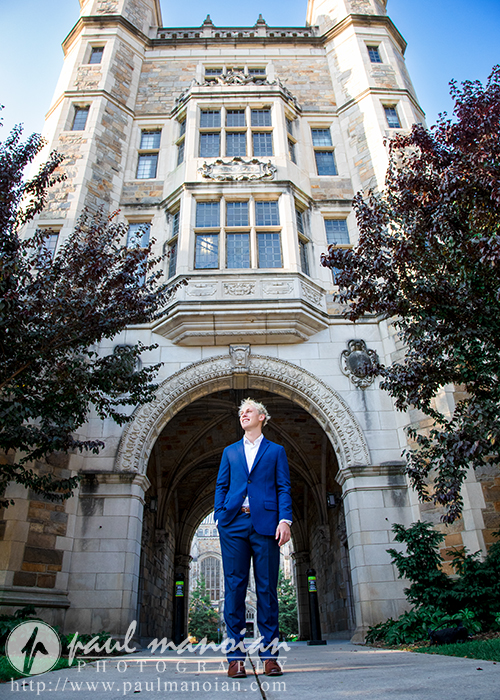 A young man in a blue suit stands confidently under a large stone archway of a historic building, sunlight filtering through trees—an elegant moment from his Ann Arbor senior portraits session.