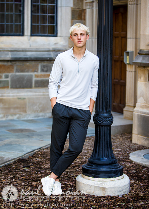 A young man with short blond hair, wearing a white long-sleeve shirt, dark pants, and white sneakers, leans against a black lamppost outside a stone building—a classic moment from an Ann Arbor senior portraits session.
