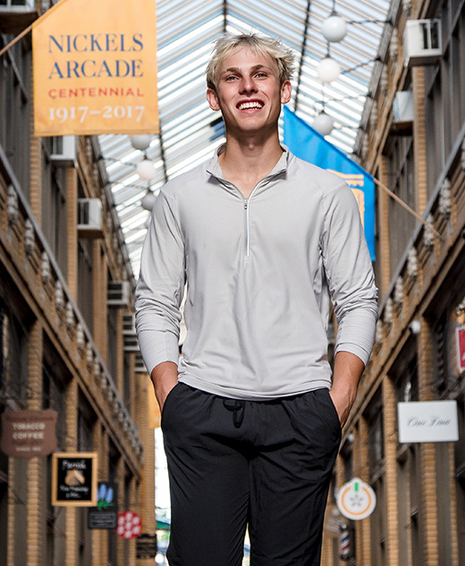 A high school senior boy with blond hair, wearing a light gray long-sleeve shirt and black pants, stands smiling in the corridor of Nickels Arcade during his Ann Arbor senior portraits session, with brick walls and shop signs visible in the background.