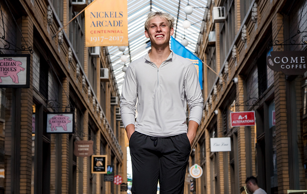 A high school senior boy with blond hair, wearing a light gray long-sleeve shirt and black pants, stands smiling in the corridor of Nickels Arcade during his Ann Arbor senior portraits session, with brick walls and shop signs visible in the background.
