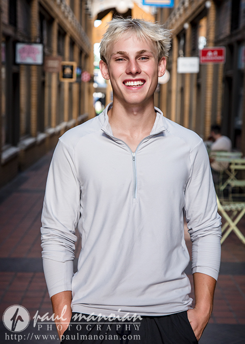 A high school senior with light blond hair smiles brightly, standing in a narrow outdoor corridor with brick walls and patio tables during his Ann Arbor senior portraits session. He wears a light gray long-sleeve shirt and has his hands in his pockets.