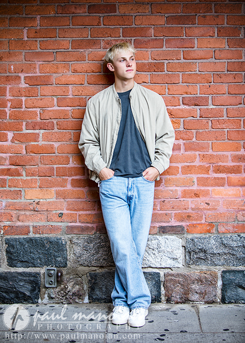 A high school senior guy with short blond hair, wearing a beige jacket, gray shirt, and blue jeans, stands casually against a red brick wall during his Ann Arbor senior portraits session, hands in his pockets as he looks thoughtfully to the side.