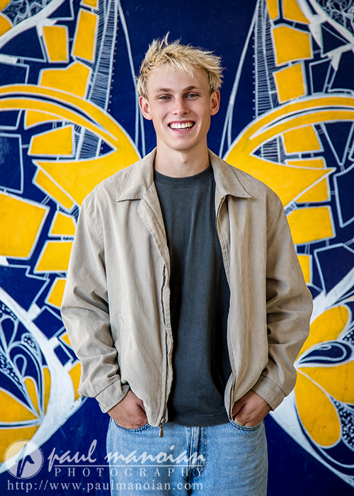 A high school senior guy with blond hair smiles while standing in front of a colorful mural during his Ann Arbor senior portraits session. He wears a beige jacket over a dark t-shirt and light blue jeans.