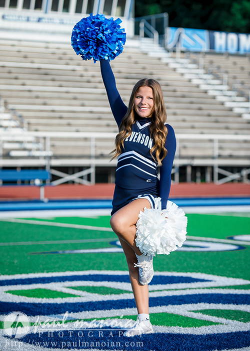 A cheerleader in a navy blue and white Stevenson High School uniform poses on the field, holding blue and white pom-poms and smiling, with empty bleachers in the background—perfect for Cheer Senior Pictures.