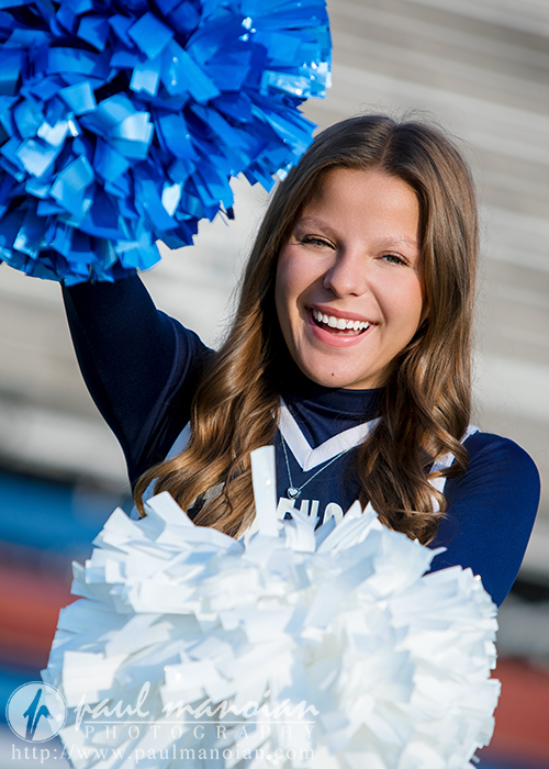 A smiling cheerleader in a navy and white uniform holds blue and white pom-poms, raising her arms enthusiastically on the field for her Stevenson High School Cheer Senior Pictures, framed by blurred stadium bleachers.