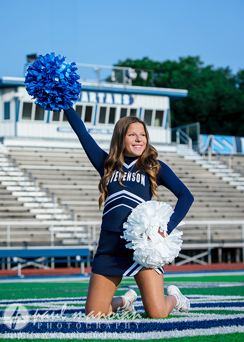 A cheerleader in a blue and white uniform kneels on the field at Stevenson High School, holding blue and white pom-poms. She is smiling brightly and raising one arm enthusiastically, with bleachers and the school building in the background.