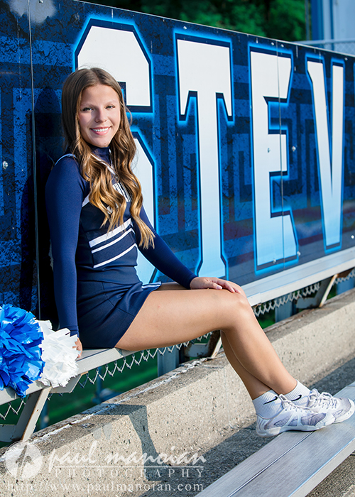 A teenage cheerleader in a navy blue uniform sits on the bleachers, holding blue and white pom-poms, smiling at the camera. Large blue and white letters spelling "STEV" are visible behind her, perfect for Cheer Senior Pictures at Stevenson High School.