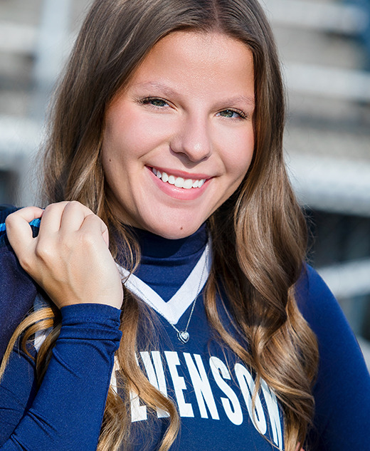 A smiling young woman with long brown hair holds a blue sports jacket over her shoulder for her cheer senior pictures. She wears a blue and white "STEVENSON" top with the number 26, standing in front of bleachers.