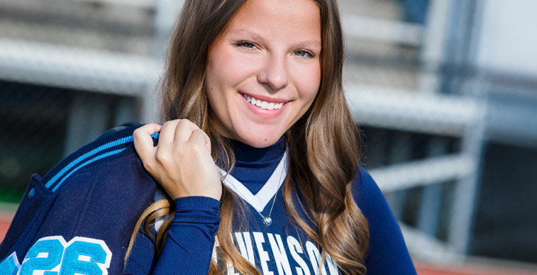 A smiling young woman with long brown hair holds a blue sports jacket over her shoulder for her cheer senior pictures. She wears a blue and white "STEVENSON" top with the number 26, standing in front of bleachers.