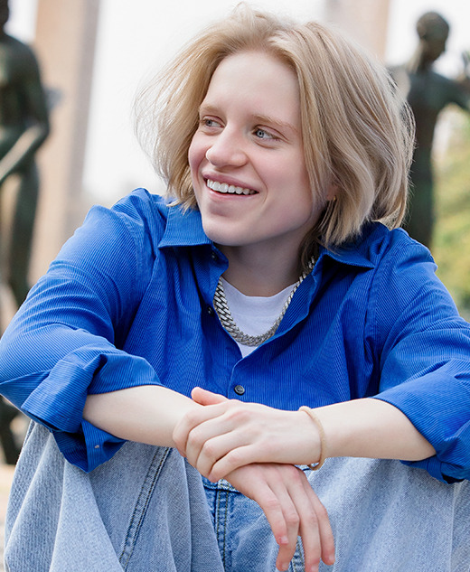 A person with light hair wearing a blue shirt and jeans sits outdoors, smiling with arms resting on their knees during a Cranbrook senior pictures session. Abstract bronze statues and greenery are visible in the background.