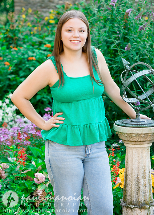 A young woman with long brown hair, wearing a green sleeveless top and light blue jeans, stands in a colorful garden during her Dearborn senior pictures session, smiling with one hand on her hip and the other resting on a stone sundial.