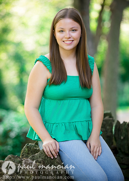 A young woman with long brown hair, wearing a green sleeveless top and light blue jeans, sits outdoors on a stone wall, smiling at the camera during her Dearborn senior pictures session. Sunlight and greenery fill the blurred background.
