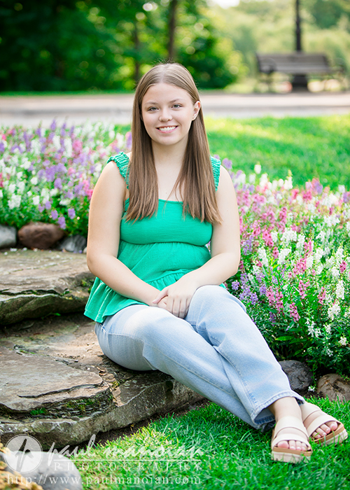 A young woman with long brown hair, wearing a green sleeveless top and light blue jeans, sits on stone steps in a garden surrounded by colorful flowers and greenery, smiling at the camera during her Dearborn senior pictures session.