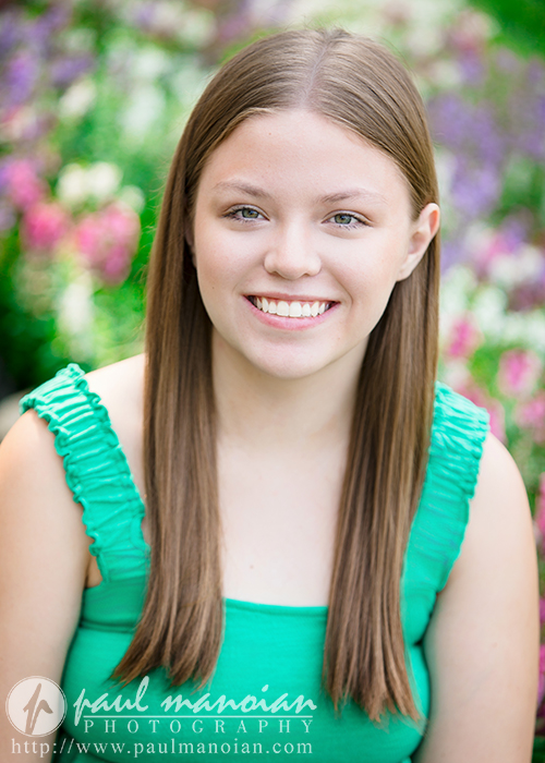 A young woman with straight brown hair, wearing a sleeveless green top, smiles at the camera during her Dearborn senior pictures session, with a blurred background of colorful flowers and greenery.