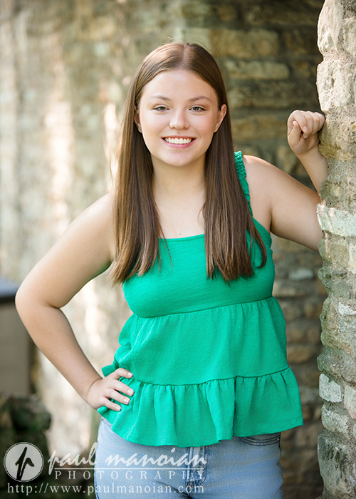 A young woman with long brown hair, wearing a sleeveless green top and jeans, smiles while standing outdoors next to a stone wall during her Dearborn senior pictures session.