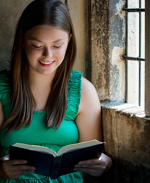 A high school senior girl in a green top and jeans sits on a stone windowsill, smiling as she reads a book, sunlight streaming beside her – a perfect moment captured during her Dearborn senior pictures session.