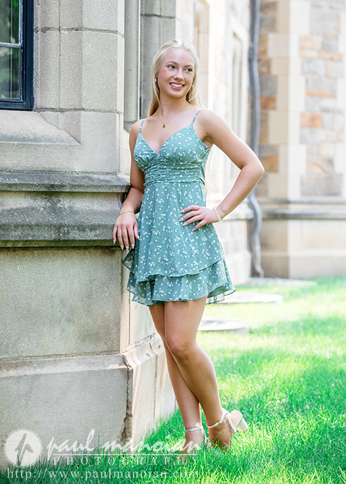 A young woman in a light blue floral dress stands smiling and leaning against a stone building on a sunny day during her Ann Arbor senior pictures session, with green grass and another stone structure in the background.
