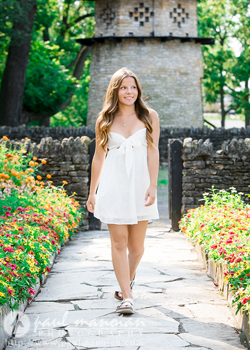 A young woman in a white dress walks along a stone path lined with colorful flowers during her Greenfield Village senior pictures session, with trees and a stone building in the background under bright daylight.