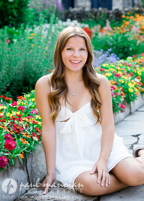 A young woman with long, wavy brown hair sits on a stone path in a garden filled with colorful flowers at her Greenfield Village senior pictures session, smiling and wearing a white dress.