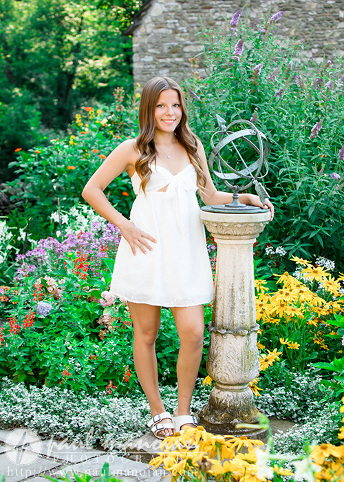 A young woman in a white dress stands in a colorful garden next to a decorative stone pedestal at her Greenfield Village senior pictures session, surrounded by blooming flowers and greenery. She smiles and poses with one hand on the pedestal.