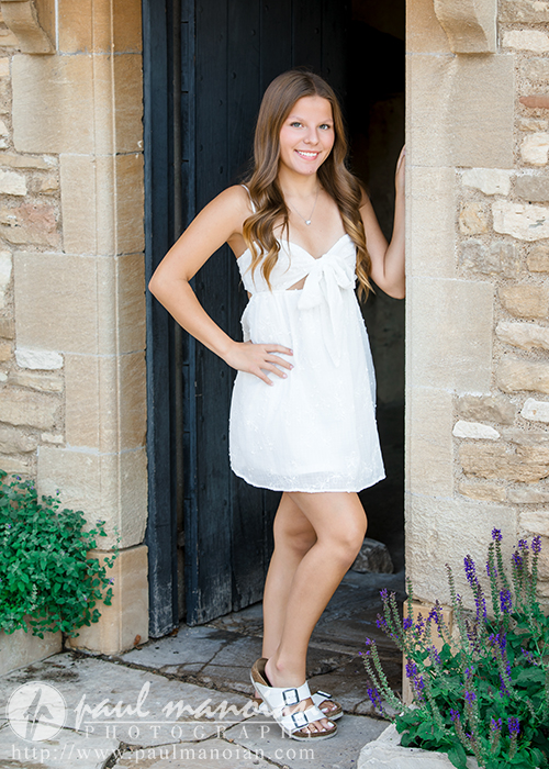 A young woman in a sleeveless white dress and sandals stands smiling in a stone doorway, hand on her hip, during her Greenfield Village senior pictures session. Purple flowers and lush greenery are visible at her feet.