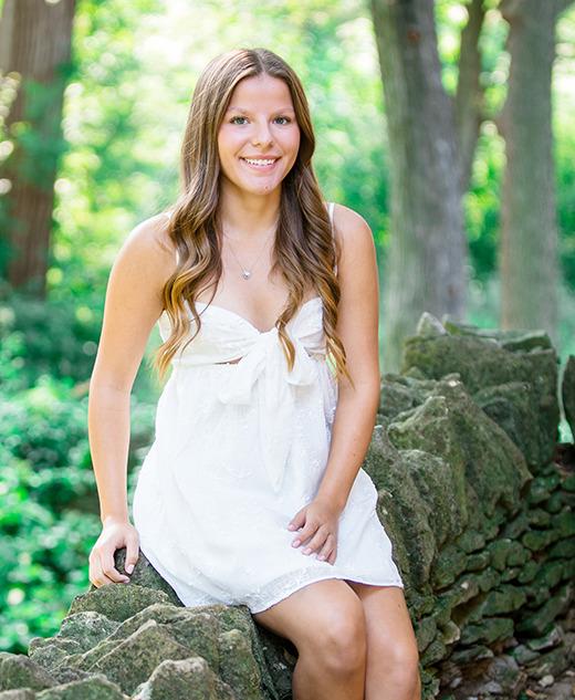 A young woman with long brown hair, wearing a sleeveless white dress, sits on a rustic stone wall in a lush, green outdoor setting at her Greenfield Village senior pictures session. Sunlight filters through the trees as she smiles at the camera.