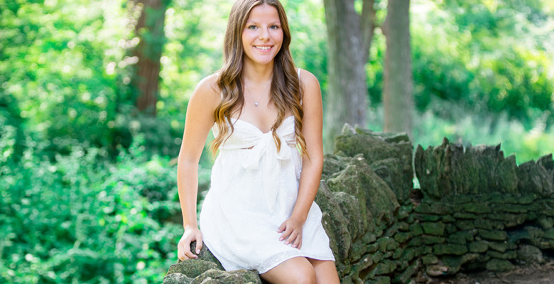 A young woman with long brown hair, wearing a sleeveless white dress, sits on a rustic stone wall in a lush, green outdoor setting at her Greenfield Village senior pictures session. Sunlight filters through the trees as she smiles at the camera.