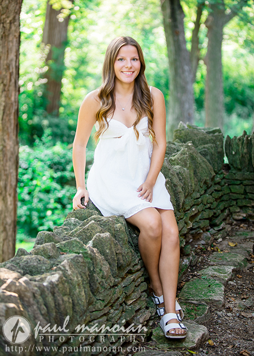 A young woman with long brown hair in a white dress sits on a stone wall during her Greenfield Village senior pictures session, smiling at the camera as sunlight filters through the lush, green trees behind her.