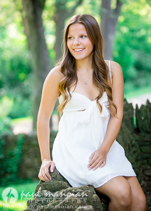 A young woman with long brown hair in a white dress sits on a stone ledge outdoors, smiling and looking to the side during her Greenfield Village senior pictures session. The lush, green background of trees is softly blurred.