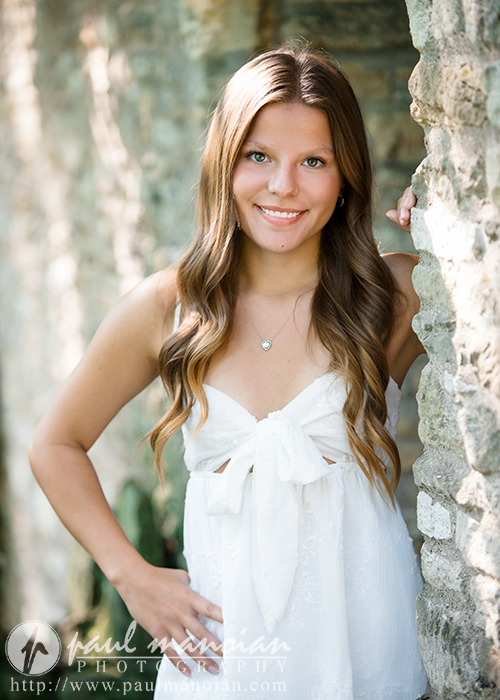 A young woman with long, wavy brown hair smiles while standing outdoors by a stone wall during her Greenfield Village senior pictures session. She is wearing a white sleeveless dress and a delicate necklace as sunlight filters through the trees in the background.