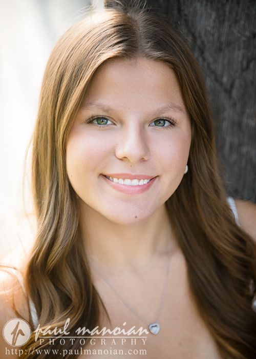 A young woman with long brown hair and green eyes smiles at the camera, standing outdoors beside a tree during her Greenfield Village senior pictures session. She wears a heart-shaped necklace and a white top, sunlight softly illuminating her face.