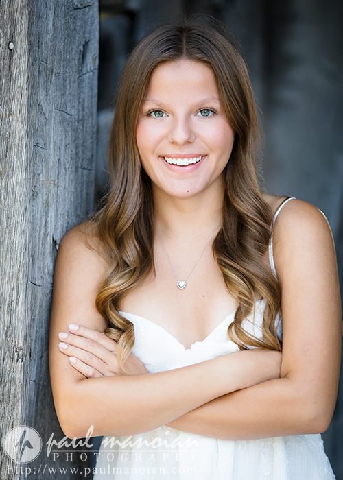 A young woman with long, wavy brown hair smiles while standing with her arms crossed in front of a rustic wooden wall during her Greenfield Village senior pictures session. She is wearing a white sleeveless dress and a silver necklace.
