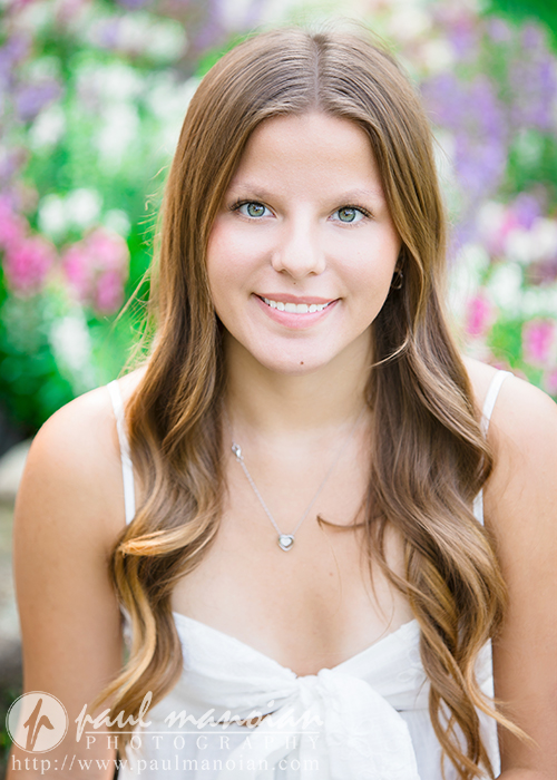 A young woman with long, wavy brown hair and light eyes smiles at the camera during her Greenfield Village senior pictures session. She wears a white sleeveless top and a necklace, sitting outdoors with colorful flowers and greenery in the background.